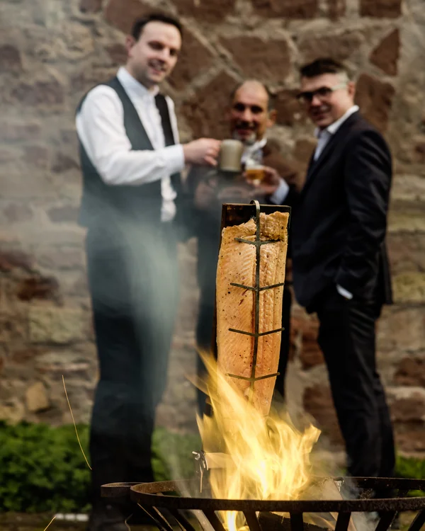 Ihr Seminar im Tagungshotel mit Rahmenprogramm Personen trinken neben einem Feuer, auf dem Lachs auf einem Holzbrett gegart wird