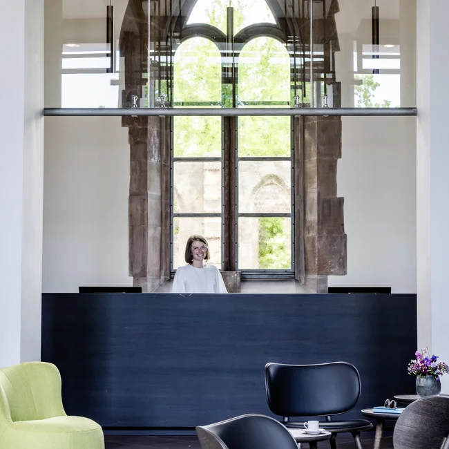 Exzellenter Lernort Kloster Hornbach Rezeption mit lächelnder Frau und modernen Stühlen in einem Raum mit gotischem Fenster