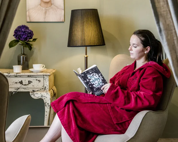 Wellness im Kloster in Deutschland Frau in rotem Bademantel liest in einem Sessel in gemütlichem Zimmer