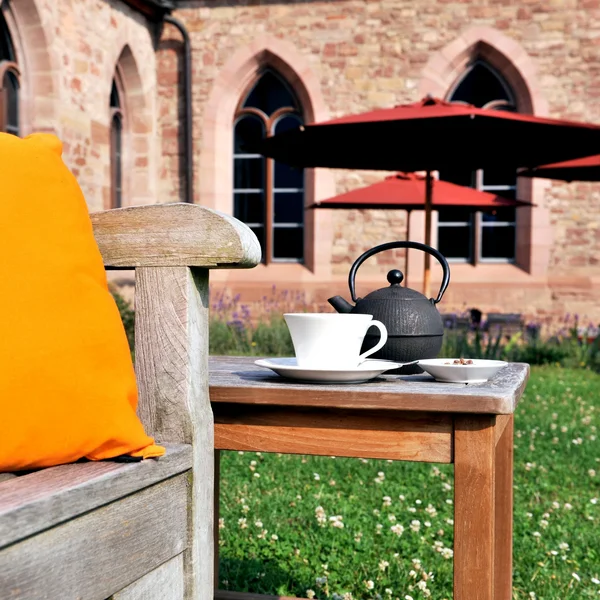 Inklusivleistungen im Kloster Hornbach und im Lösch Holztisch mit Teekanne, Tasse und orangefarbenem Kissen im Garten vor einem Steinbau