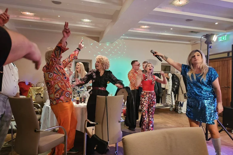 ABBA-Dinner – A Night to Remember Menschen feiern und singen mit Mikrofon auf einer Party in bunten Outfits