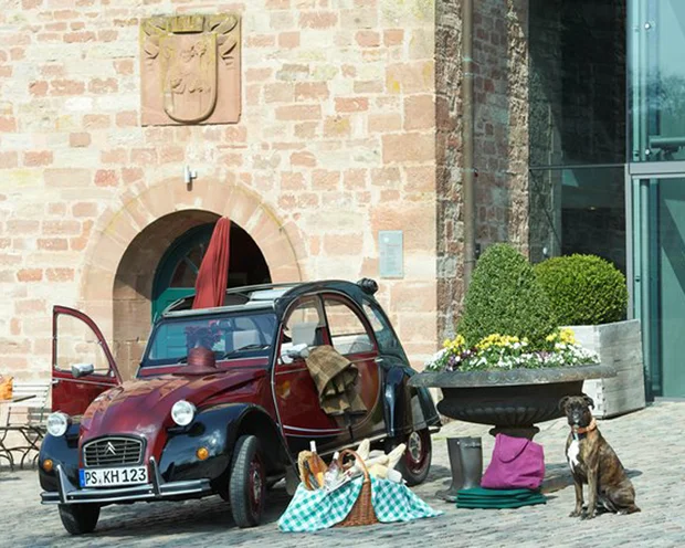 Ausflugstipps für Ihren Urlaub in der Pfalz Rotes und schwarzes Vintage-Auto mit Picknickkorb und sitzendem Hund vor altem Gebäude